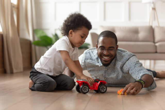 Smiling Black father and young child playing with toy cars on the floor at home